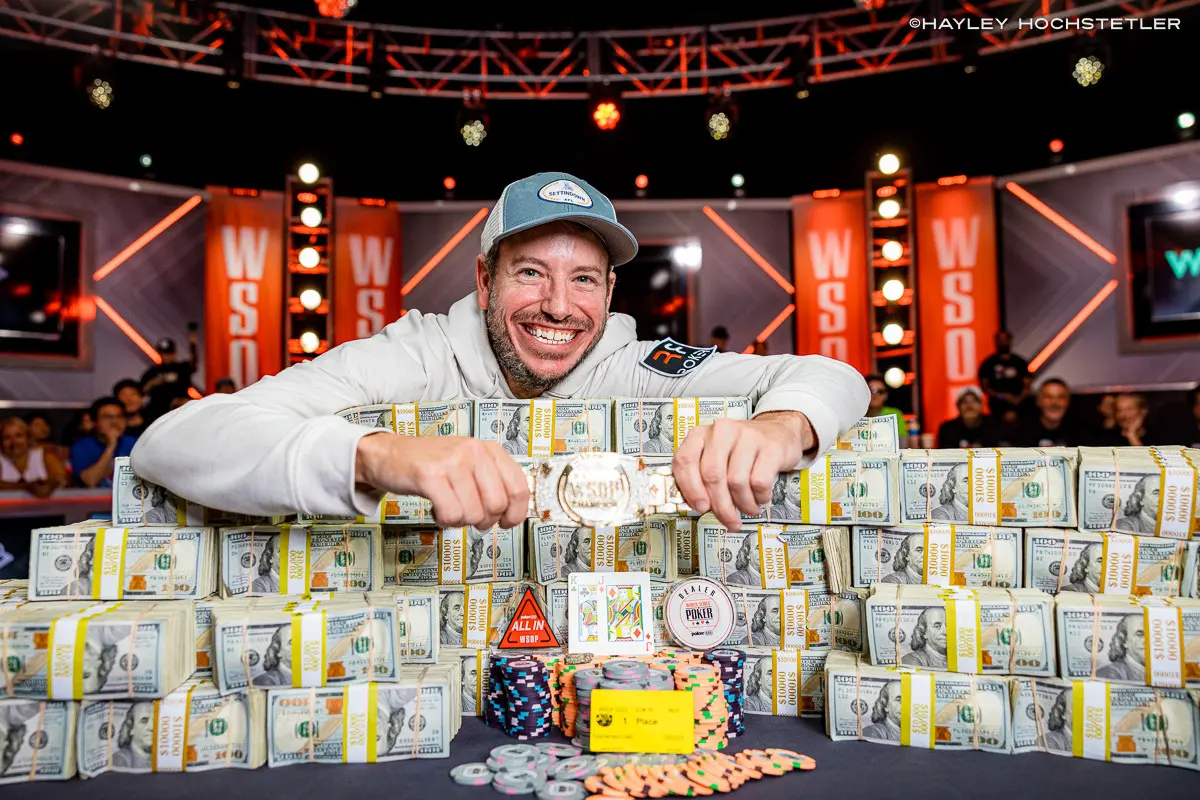 Smiling poker champion displaying trophy with stacks of cash and chips behind.