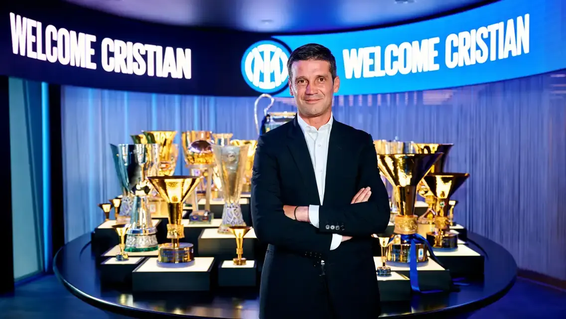 Cristian Chivu in a suit standing in front of an Inter Milan trophy display and 'Welcome Cristian' screens